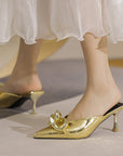 Gold high-heeled shoes with a decorative bow worn by a person in a white skirt.