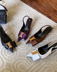 Four pairs of high-heeled shoes in black, purple, brown, and white on a textured surface.