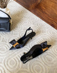 Black high-heeled shoes with gold accents on a textured surface next to a black handbag.