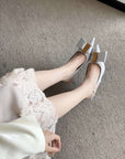 White high-heeled shoes with gold accents worn by a person in a white dress on a gray carpet.