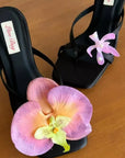 Black high-heeled sandals with pink orchid decorations on a wooden surface