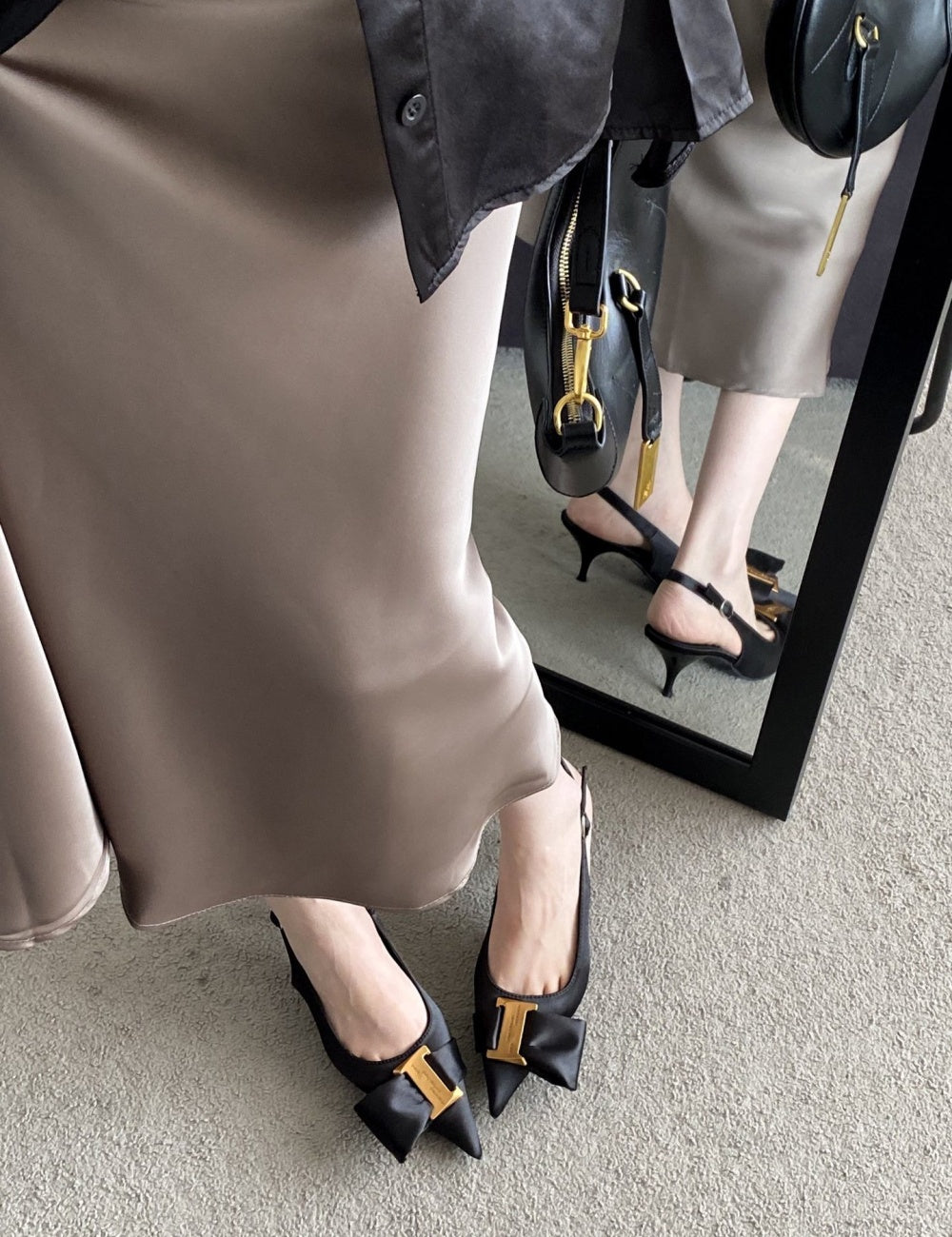 Person wearing a beige skirt and black high-heeled shoes with gold accents, standing in front of a mirror.
