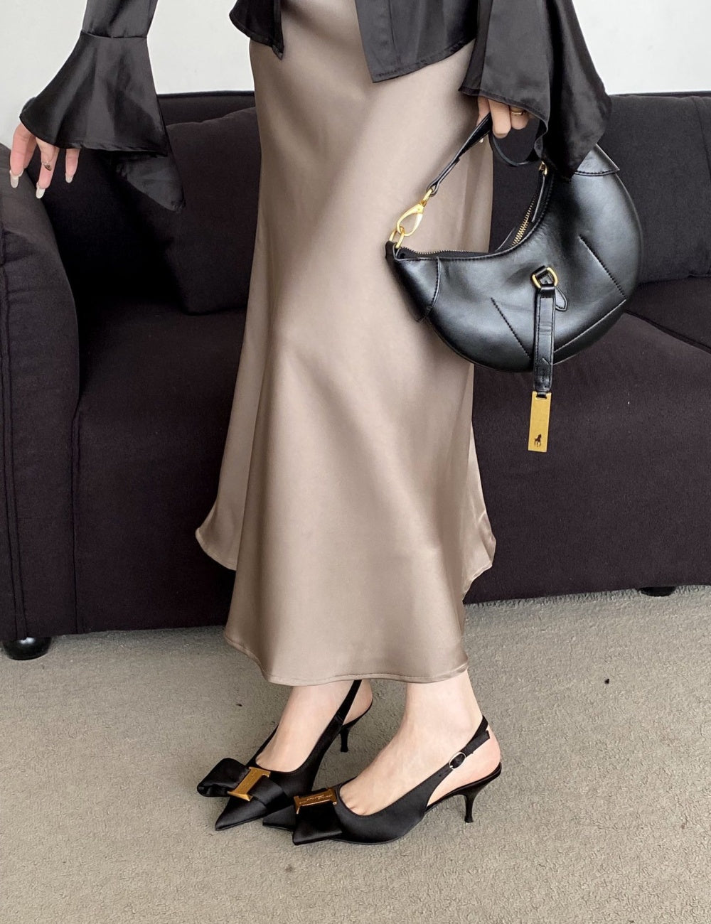Person wearing a beige skirt, black top, and black high-heeled shoes, holding a black handbag.
