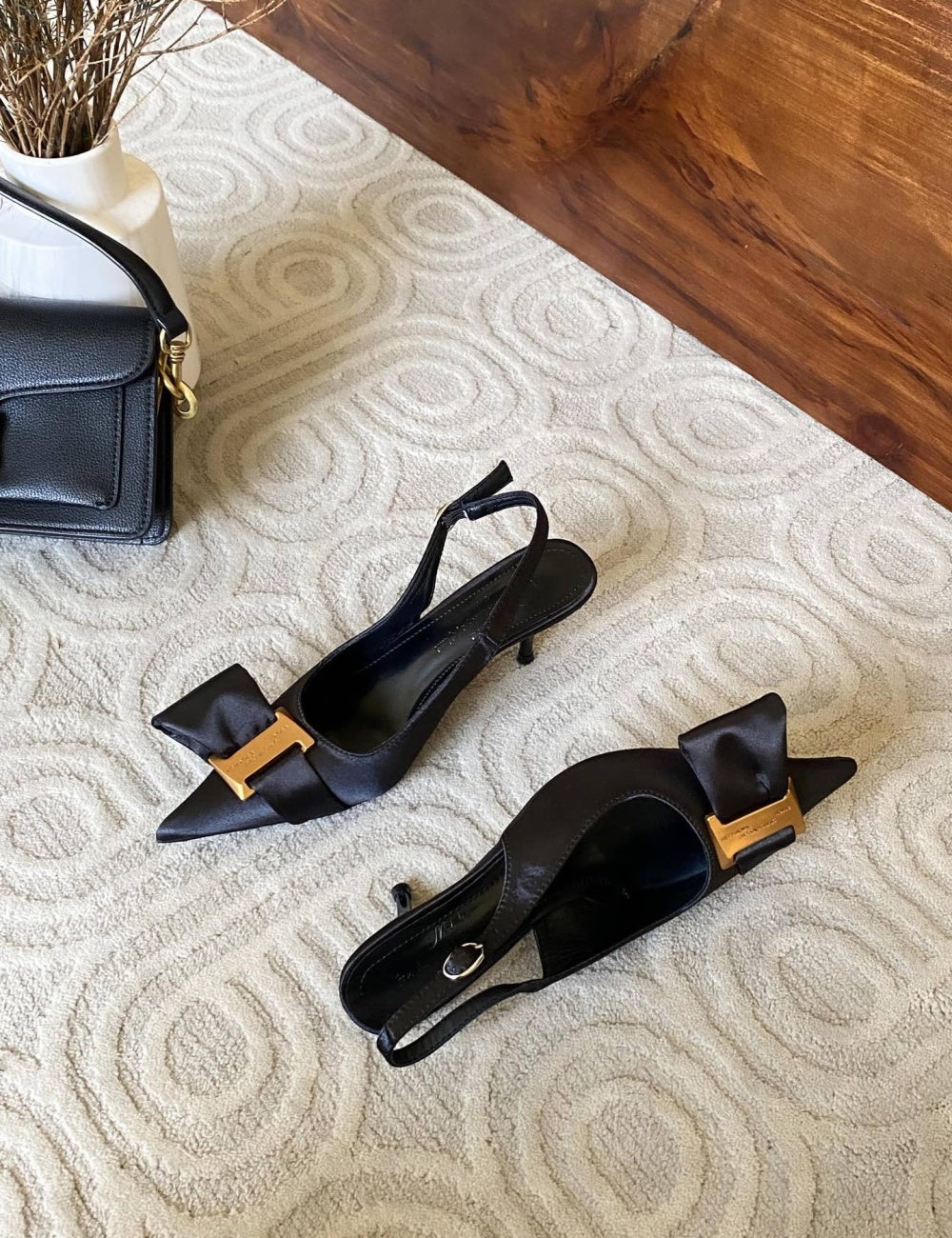 Black high-heeled shoes with gold accents on a textured surface next to a black handbag.