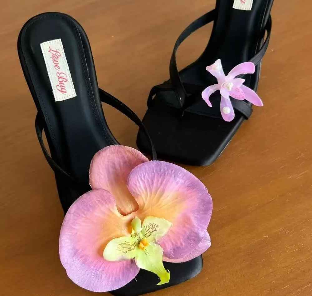 Black high-heeled sandals with pink orchid decorations on a wooden surface