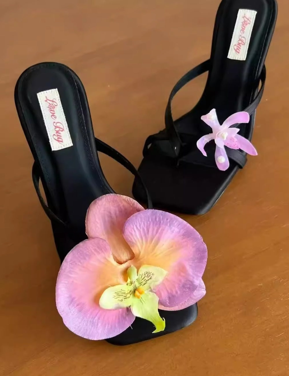 Black high-heeled sandals with pink orchids on a wooden surface